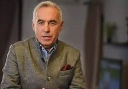 Călin Georgescu vorbește serios într-un interviu, îmbrăcat într-un sacou gri de lână, cu o eșarfă burgundy în buzunar, într-un decor interior