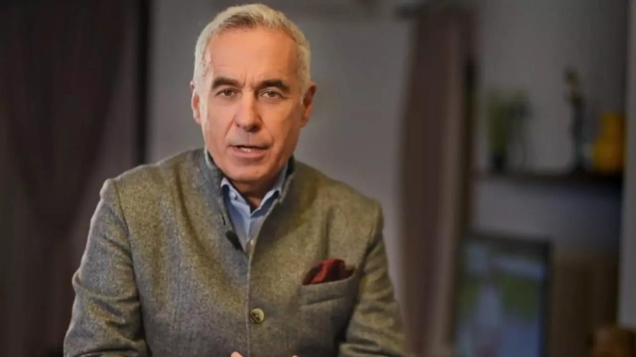 Călin Georgescu vorbește serios într-un interviu, îmbrăcat într-un sacou gri de lână, cu o eșarfă burgundy în buzunar, într-un decor interior