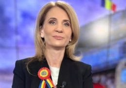 Anca Alexandrescu, prezentatoarea emisiunii Culisele Statului Paralel, zâmbind în timp ce poartă o cocardă tricoloră, reprezentând demnitatea și patriotismul în mass-media românească.