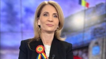 Știri de azi | Încercare disperată de intimidare la adresa televiziunii poporului, atac fără precedent: procurorul soroșist ar vrea să o citeze pe Anca Alexandrescu la Parchet – “Sunt pregătită cu demnitate, cu fruntea sus” - Știri de azi | Anca Alexandrescu, jurnalista Realitatea PLUS, surâzând în studio, purtând o rosetă tricoloră.