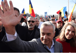 Călin Georgescu salută susținătorii în timpul unui miting, cu multitudine de steaguri românești în fundal